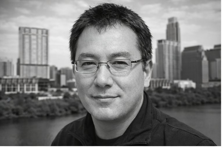 Black-and-white portrait of John Kim Austin with downtown Austin towers rising across the river behind him